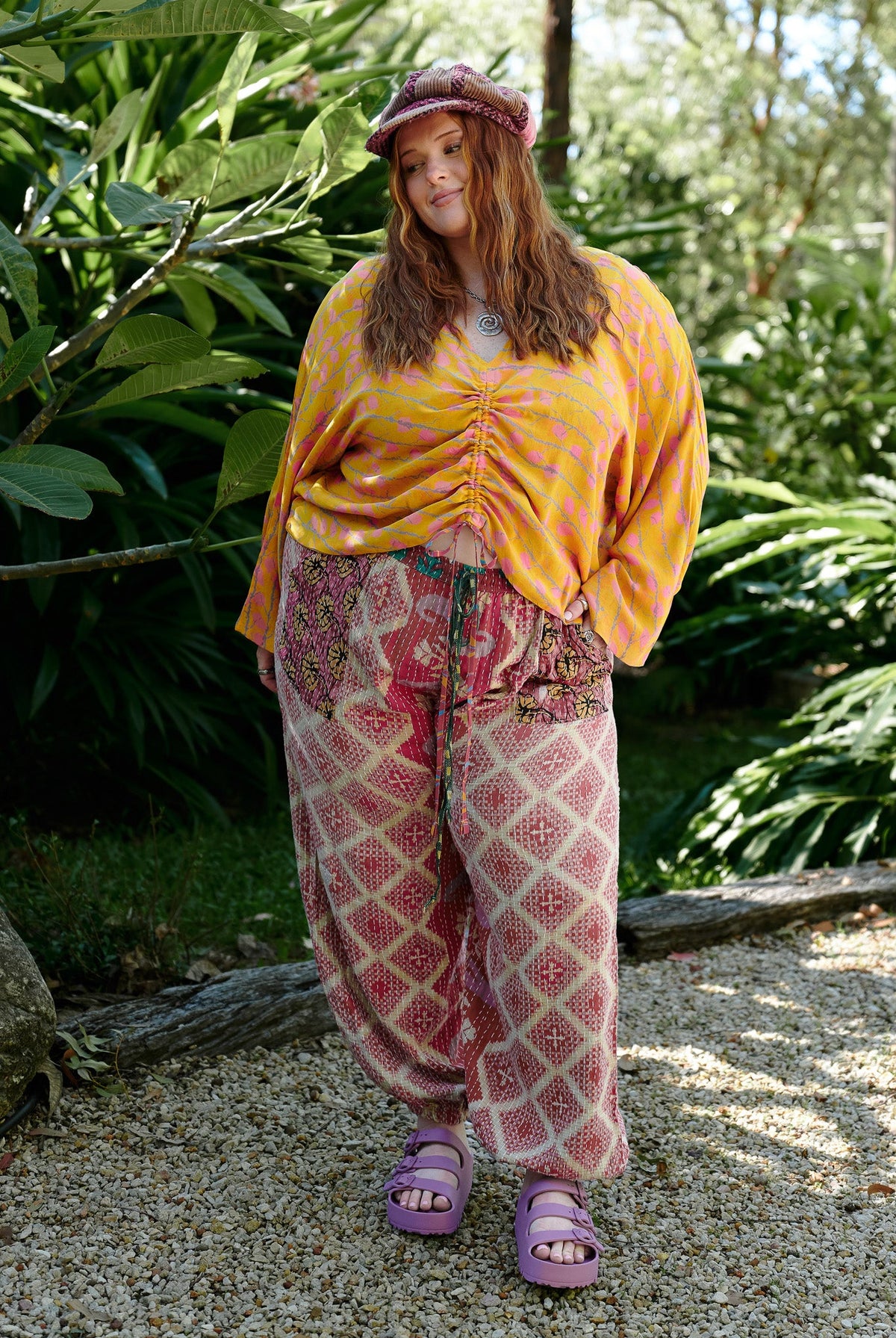 Person wearing a yellow top and patterned pants standing in a lush green forest.