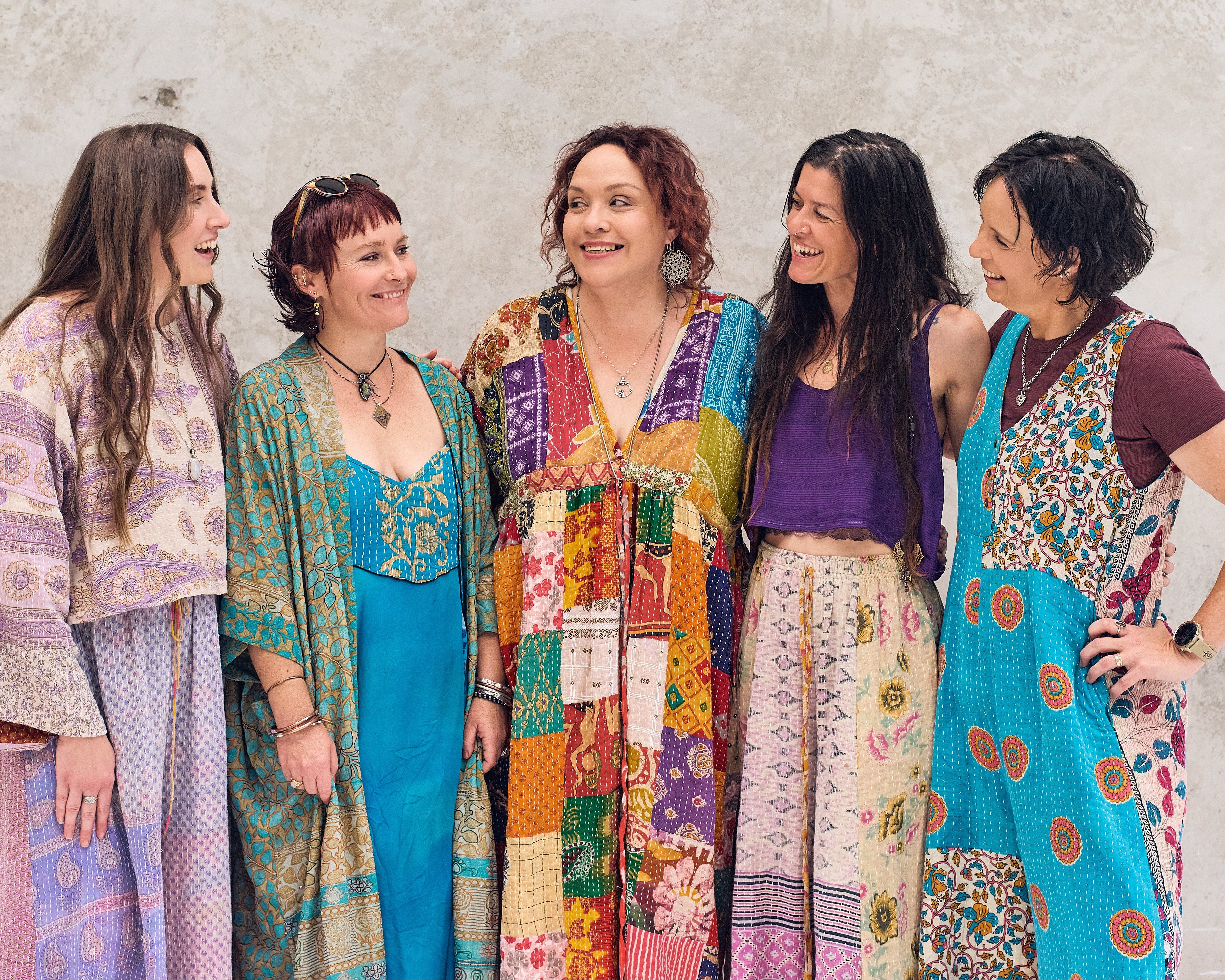 Five women wearing colorful, patterned dresses standing against a textured wall.