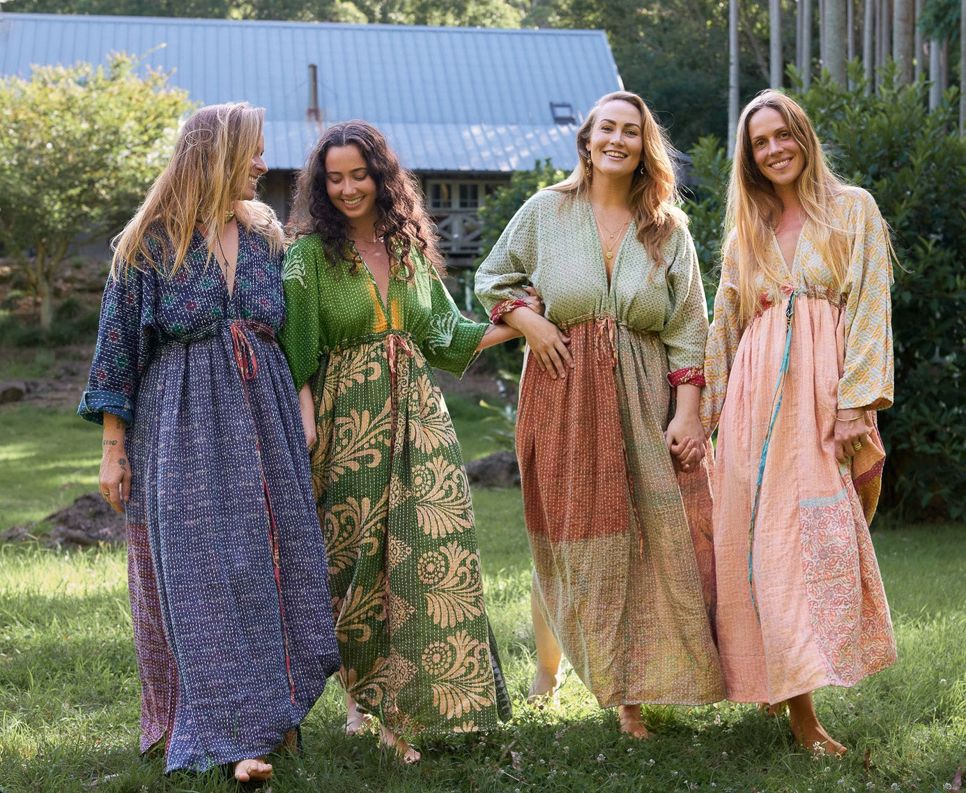Four women in long, patterned dresses standing in a grassy area with a rustic building in the background.
