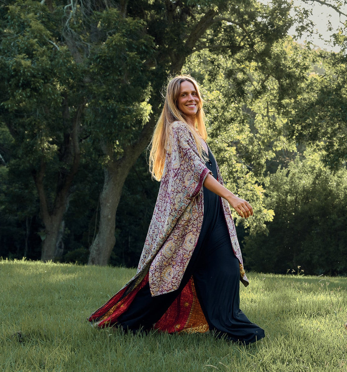 A women in flowing dress with floral patterns walking in a grassy field with trees in the background.