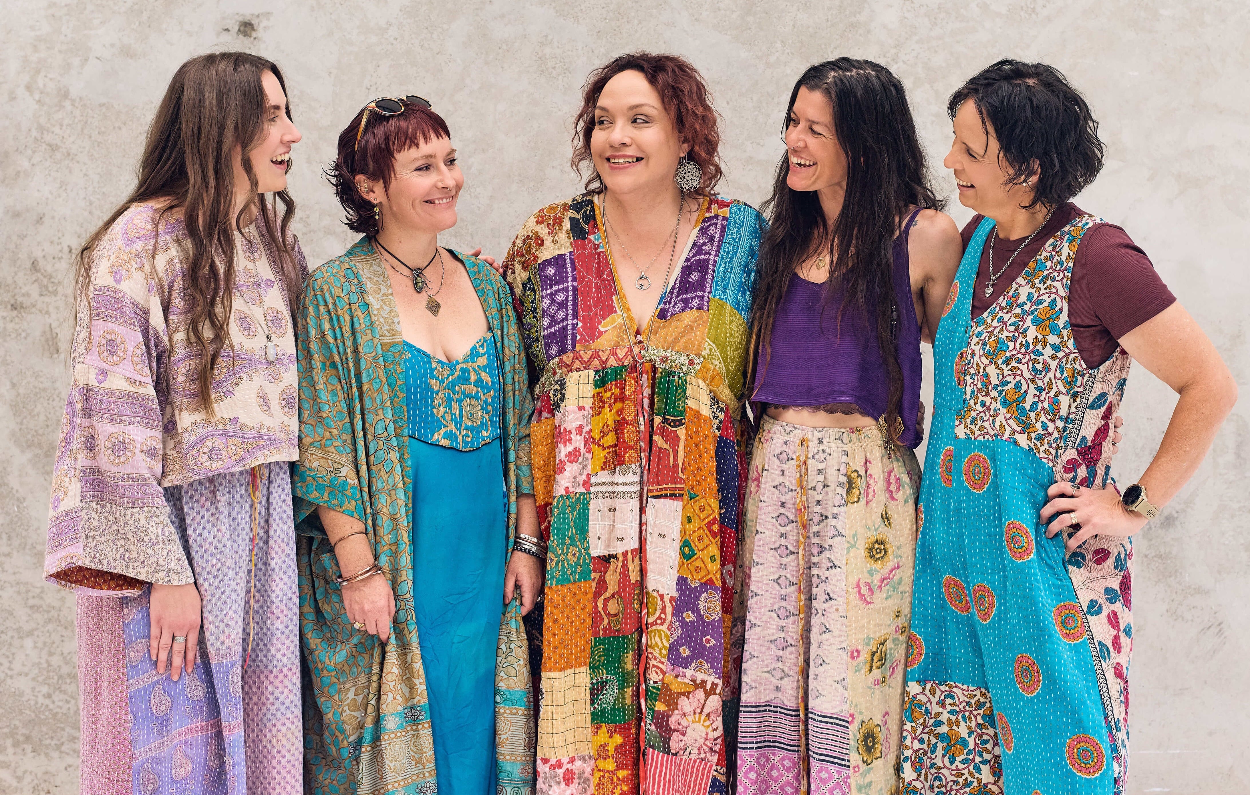 Five women wearing colorful, patterned dresses standing against a textured wall.