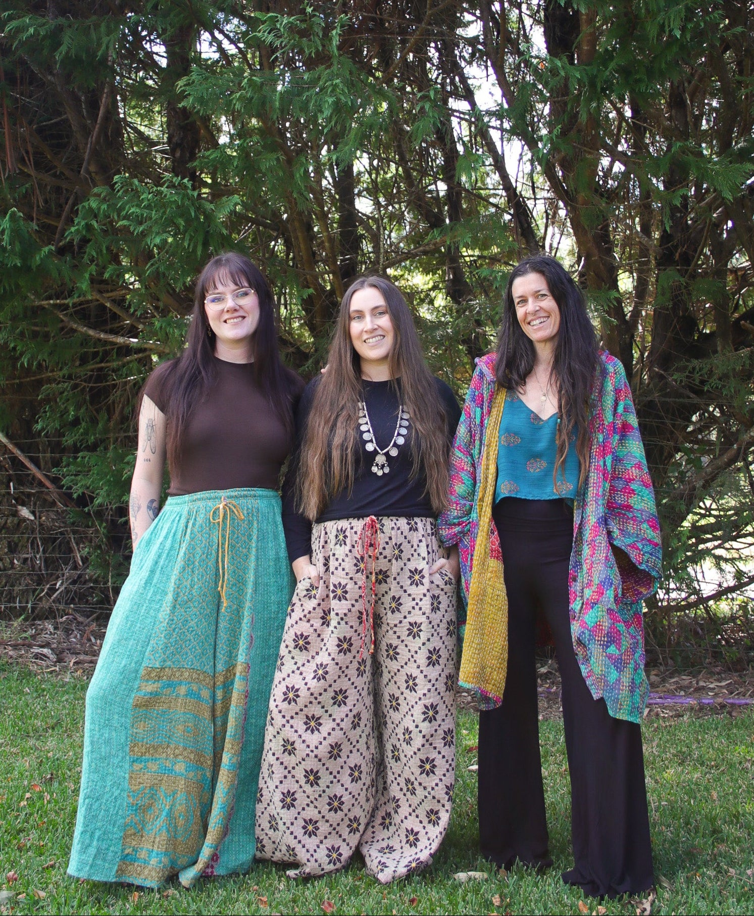 Three women standing outdoors in front of trees, wearing colorful outfits.