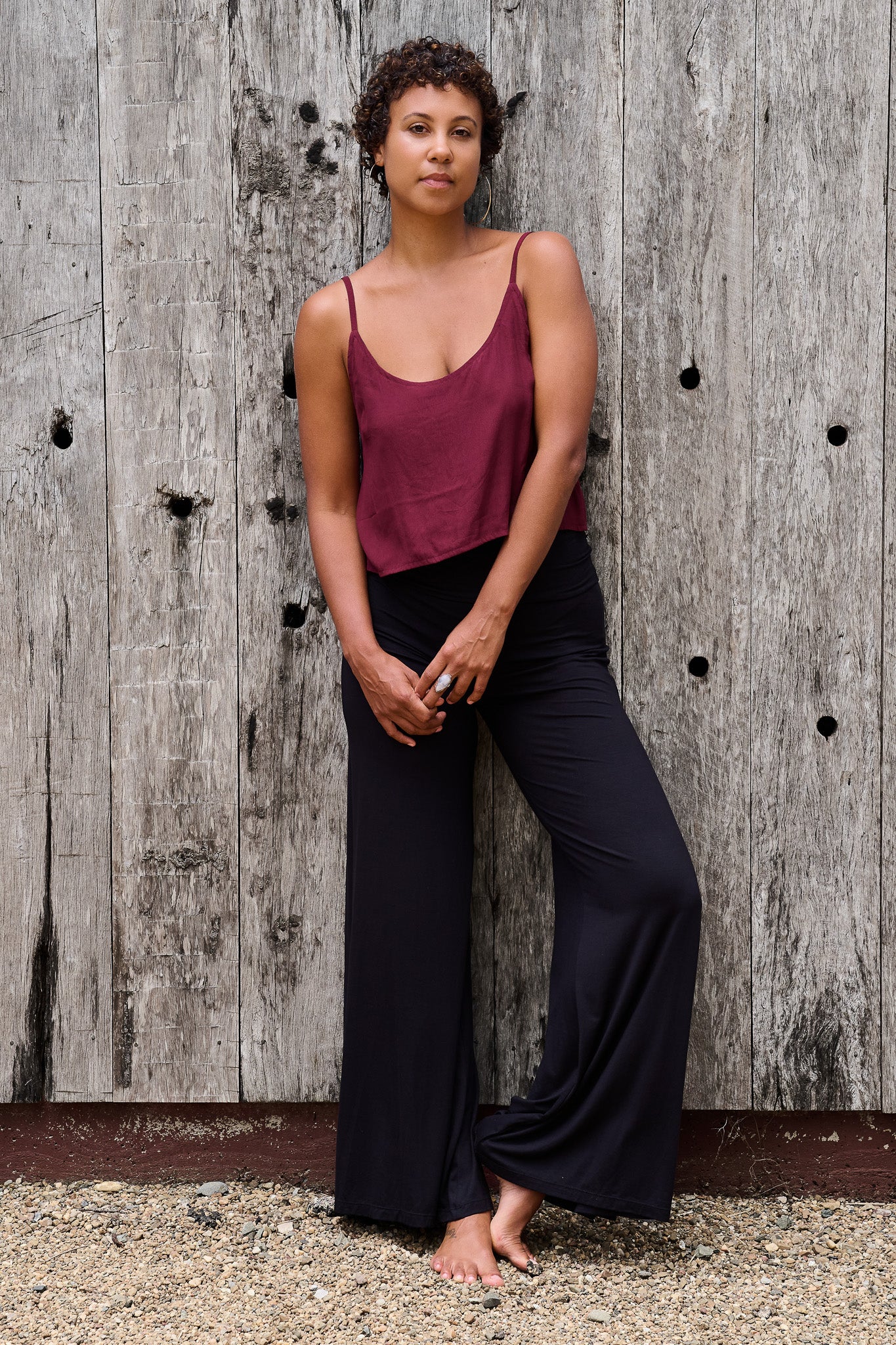 Woman wearing a burgundy tank top and black pants against a wooden wall.