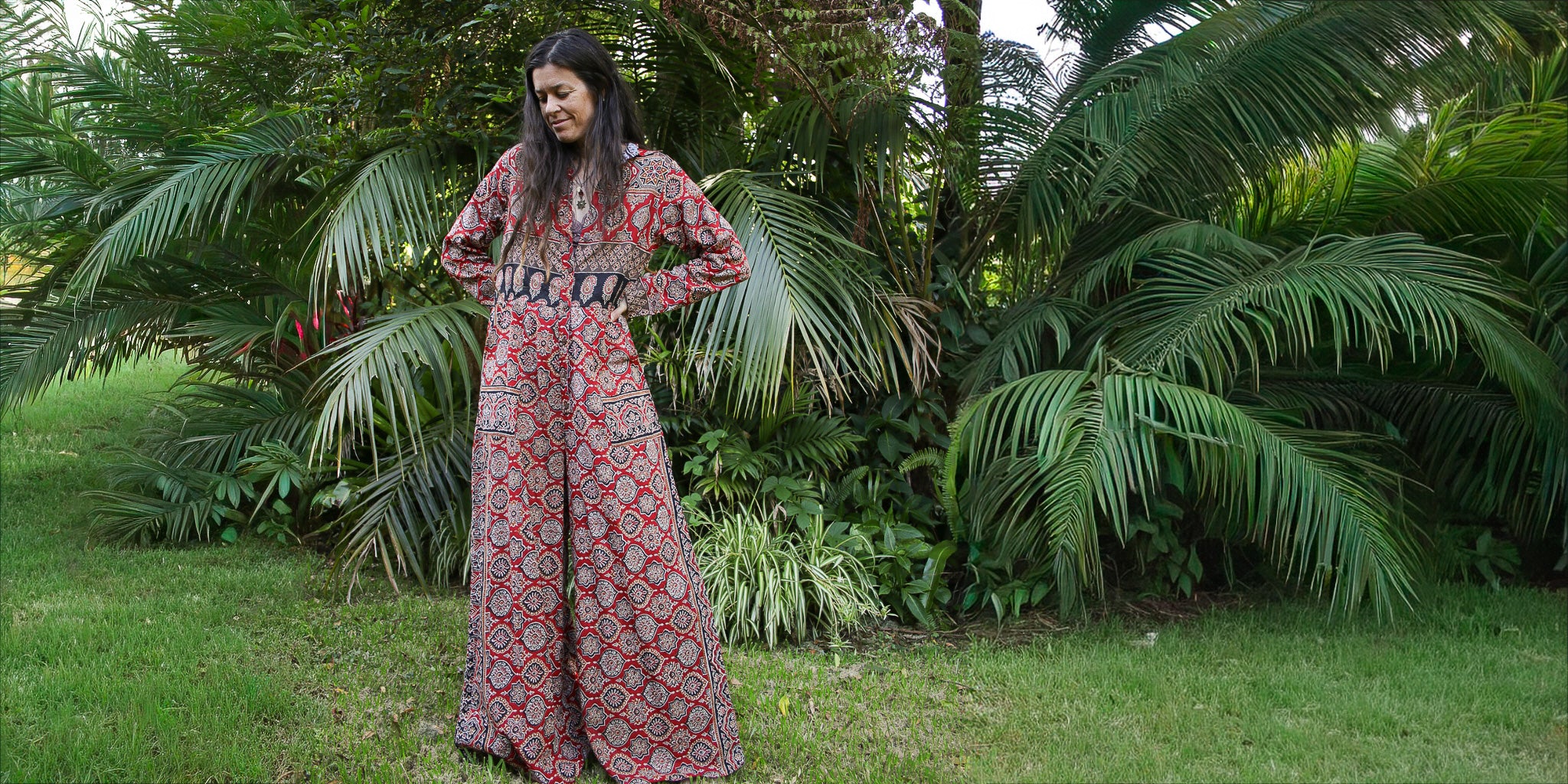 Woman in a patterned jumpsuit standing in a lush green garden with palm trees.