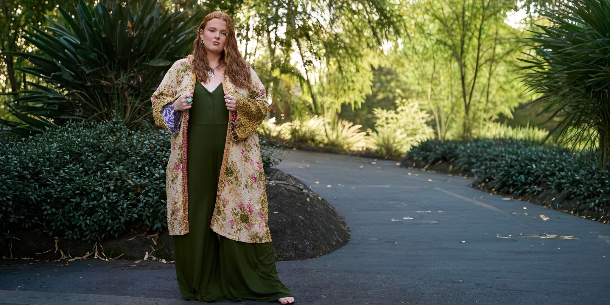 Woman in a floral dress and green long coat standing on a path in a park.