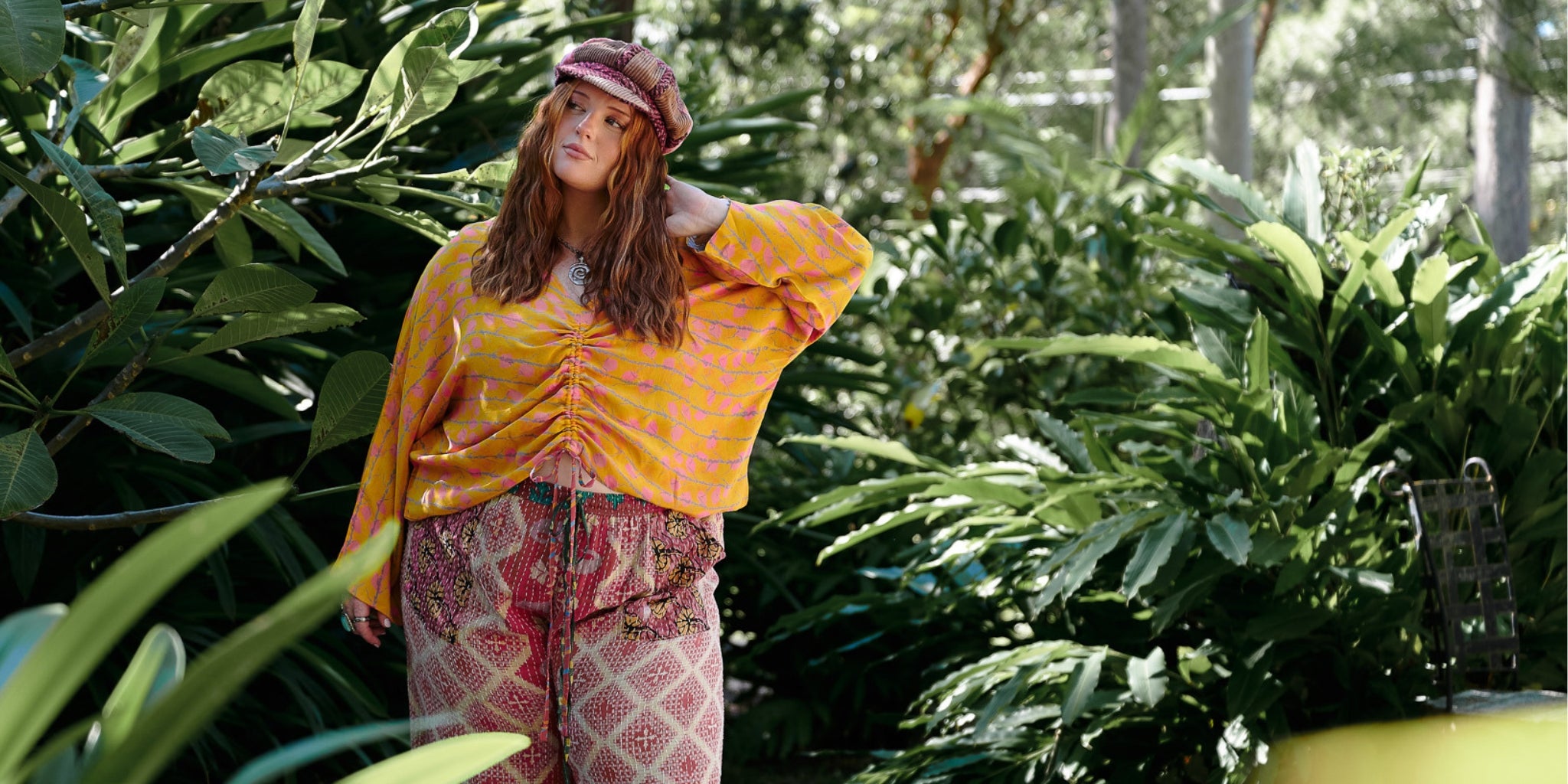 Person wearing a yellow top and patterned pants standing in a lush green forest.