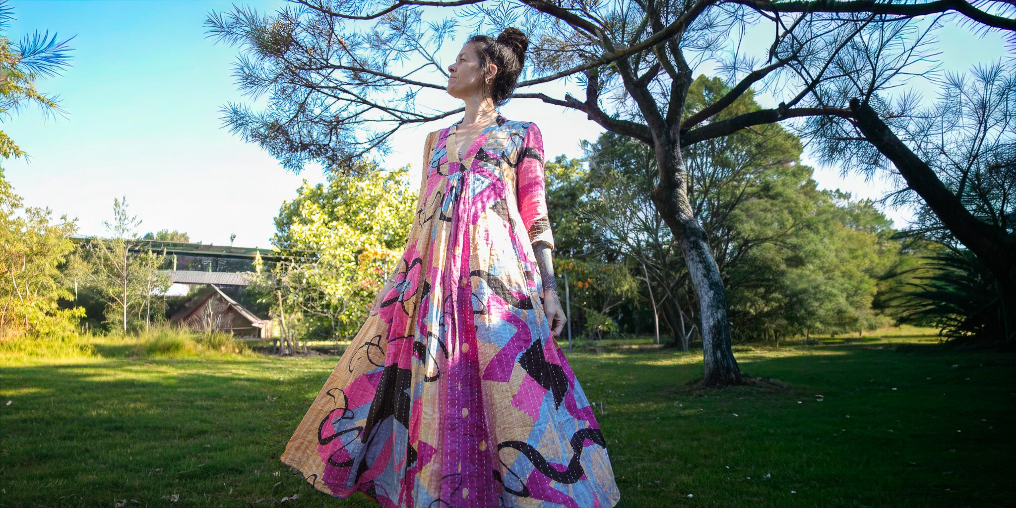 Woman in a colorful dress standing in a park with trees and a bridge in the background