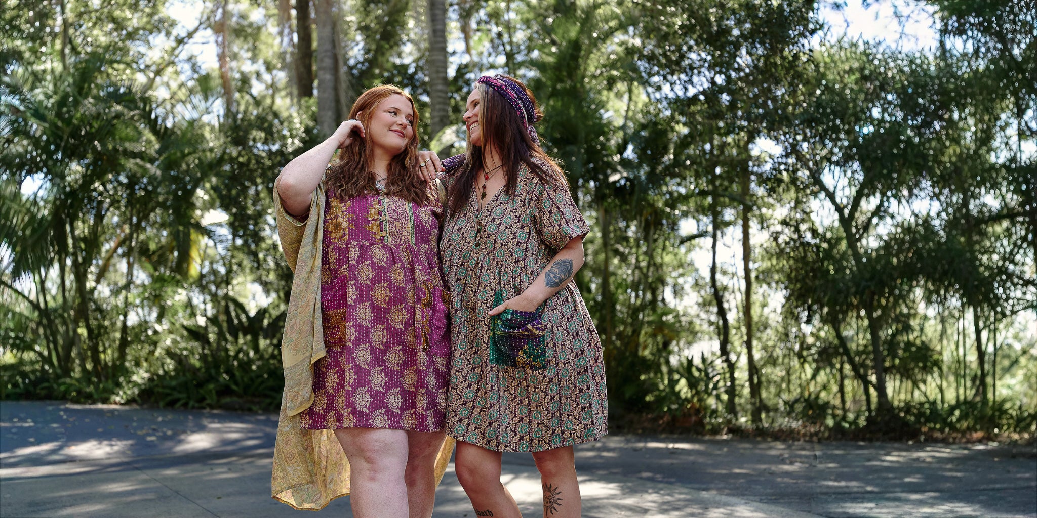 Two women standing outdoors in a forested area, wearing patterned dresses.