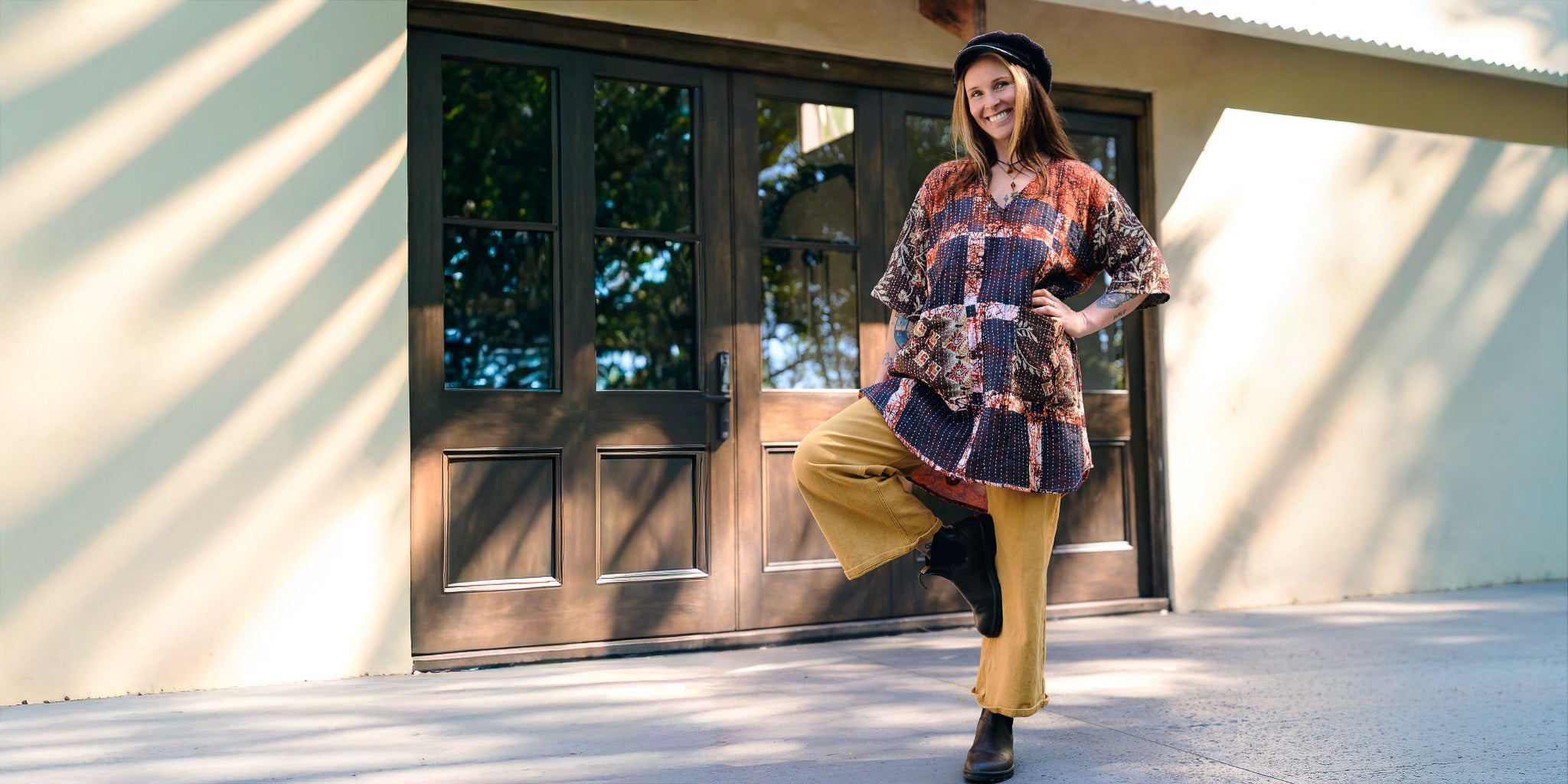 Woman in a patterned shirt and yellow pants standing in front of a wooden door.