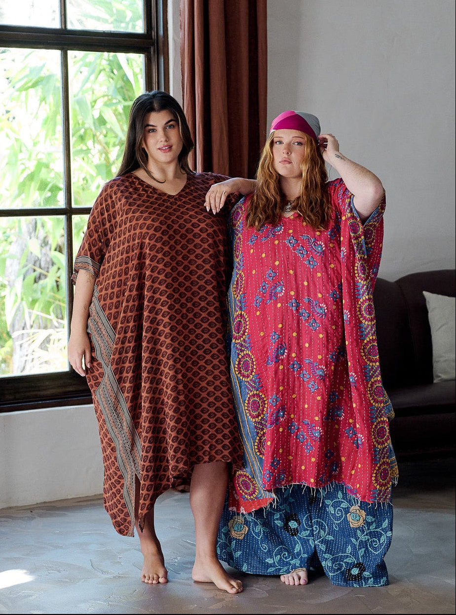 Two women standing in a room with a large window, one wearing a patterned dress and the other in a colorful outfit.