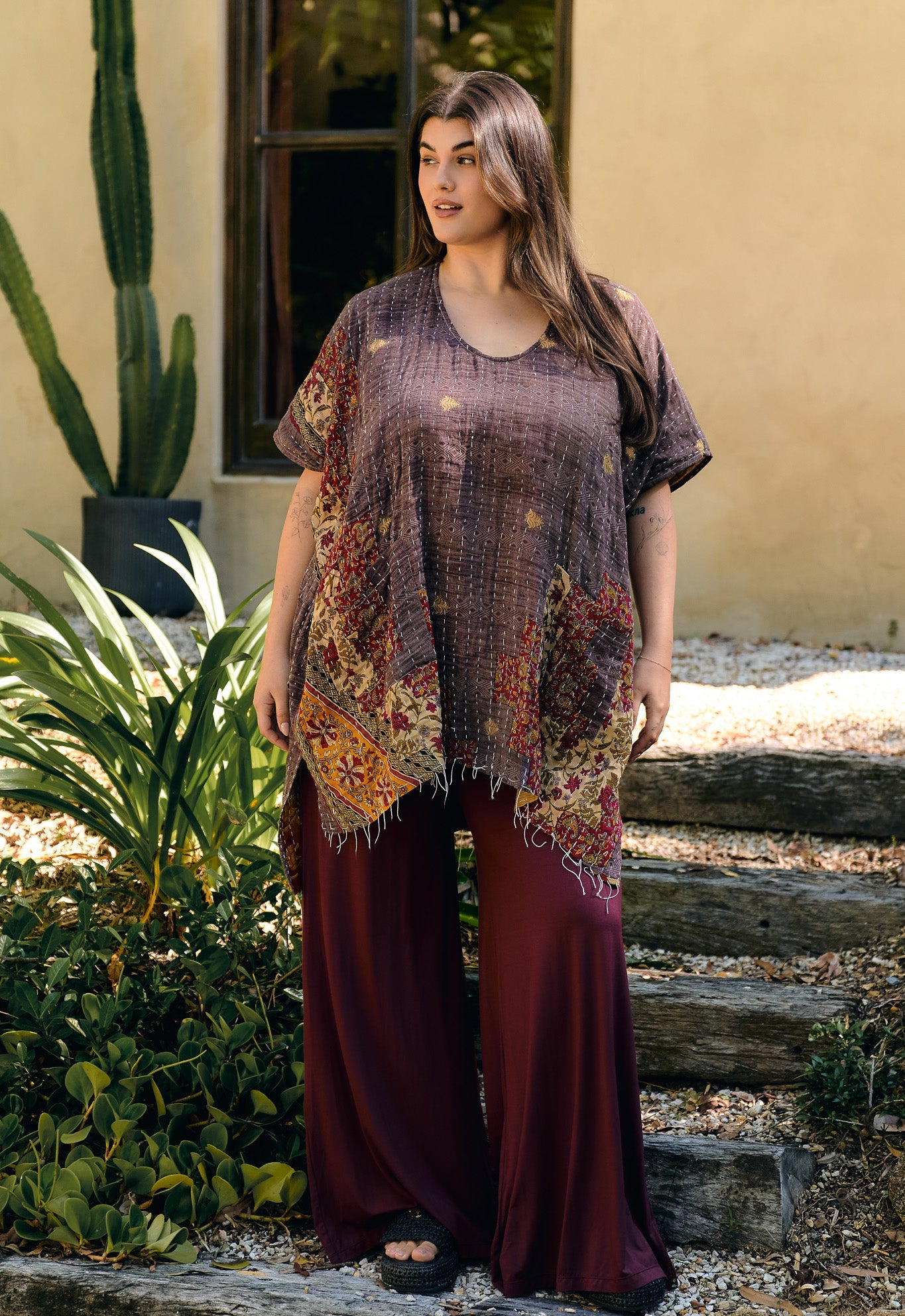 Woman wearing a patterned top and maroon pants standing outdoors with plants and a building in the background.