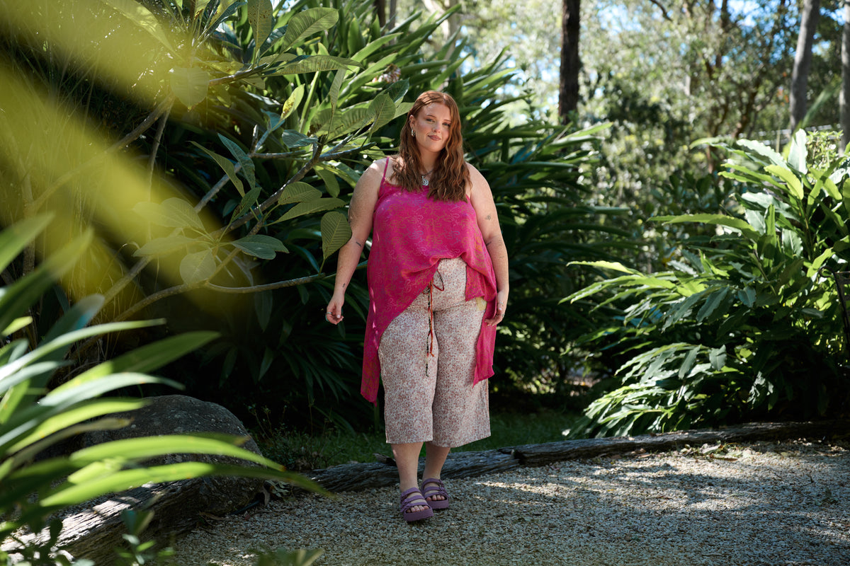 A woman walking on a gravel path surrounded by lush green tropical plants, wearing a bright pink sleeveless top, a matching pink wrap, and light beige patterned pants.