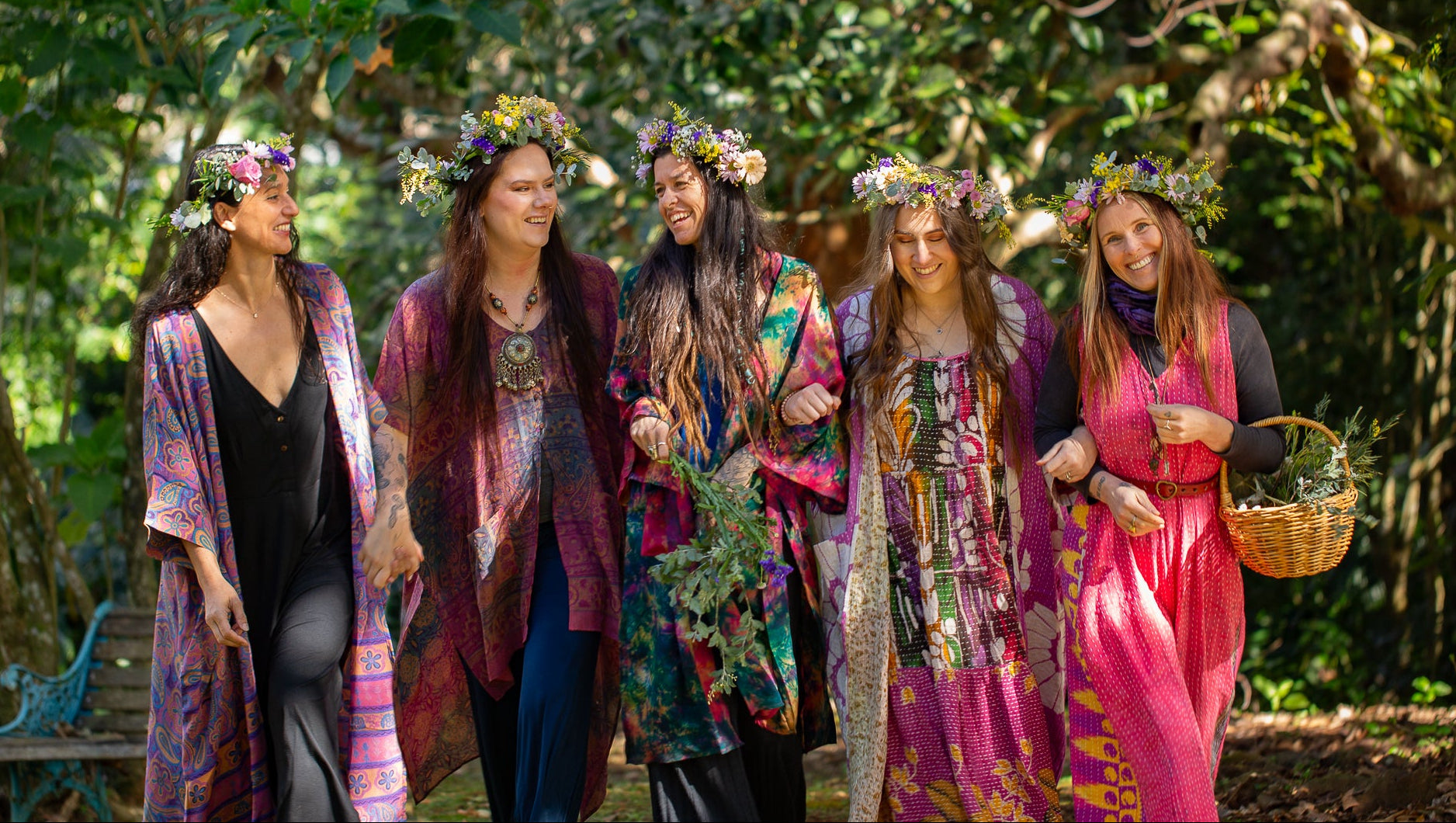 Five women wearing colorful bohemian patterned robes and flower crowns walk together in a lush green forest, with one holding a woven basket.