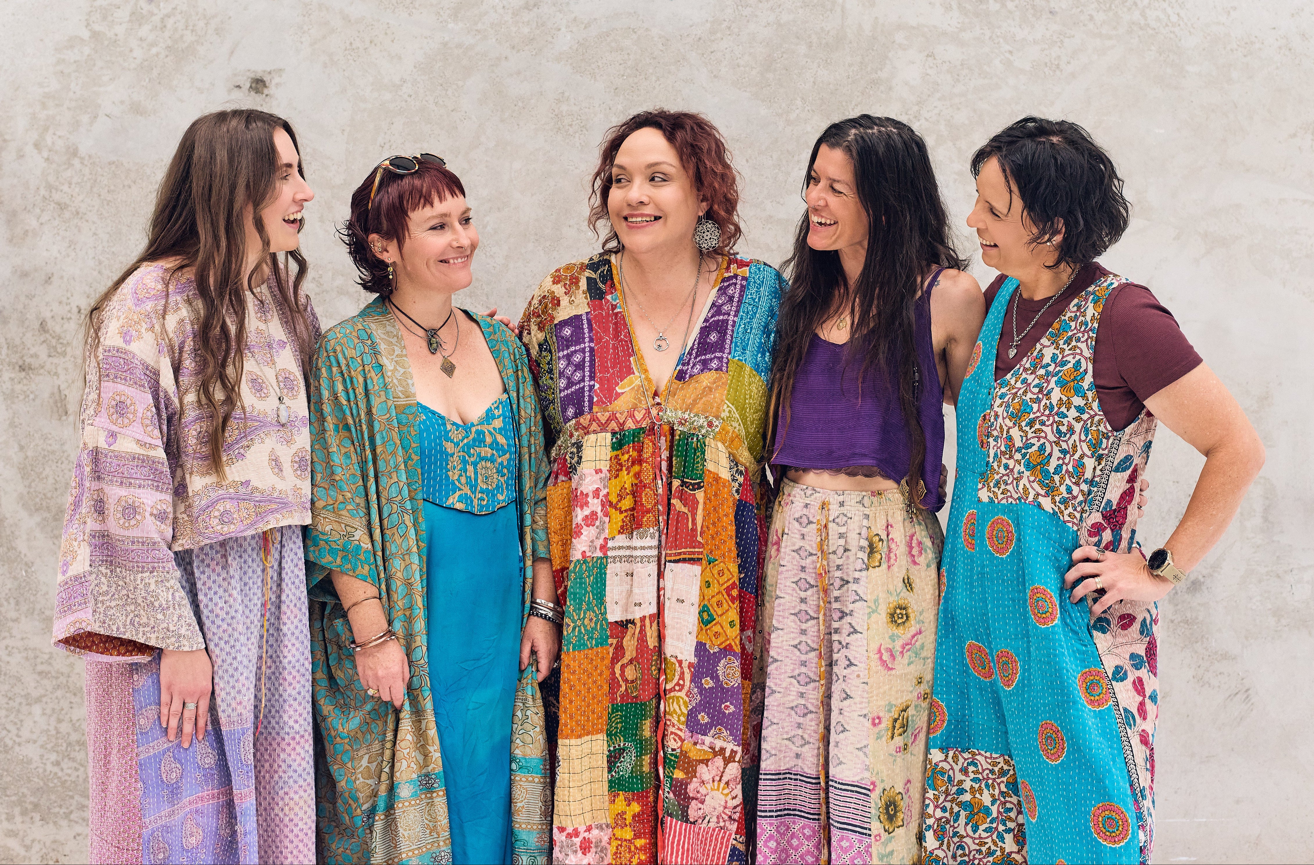 Five women wearing colorful, patterned dresses standing against a textured wall.