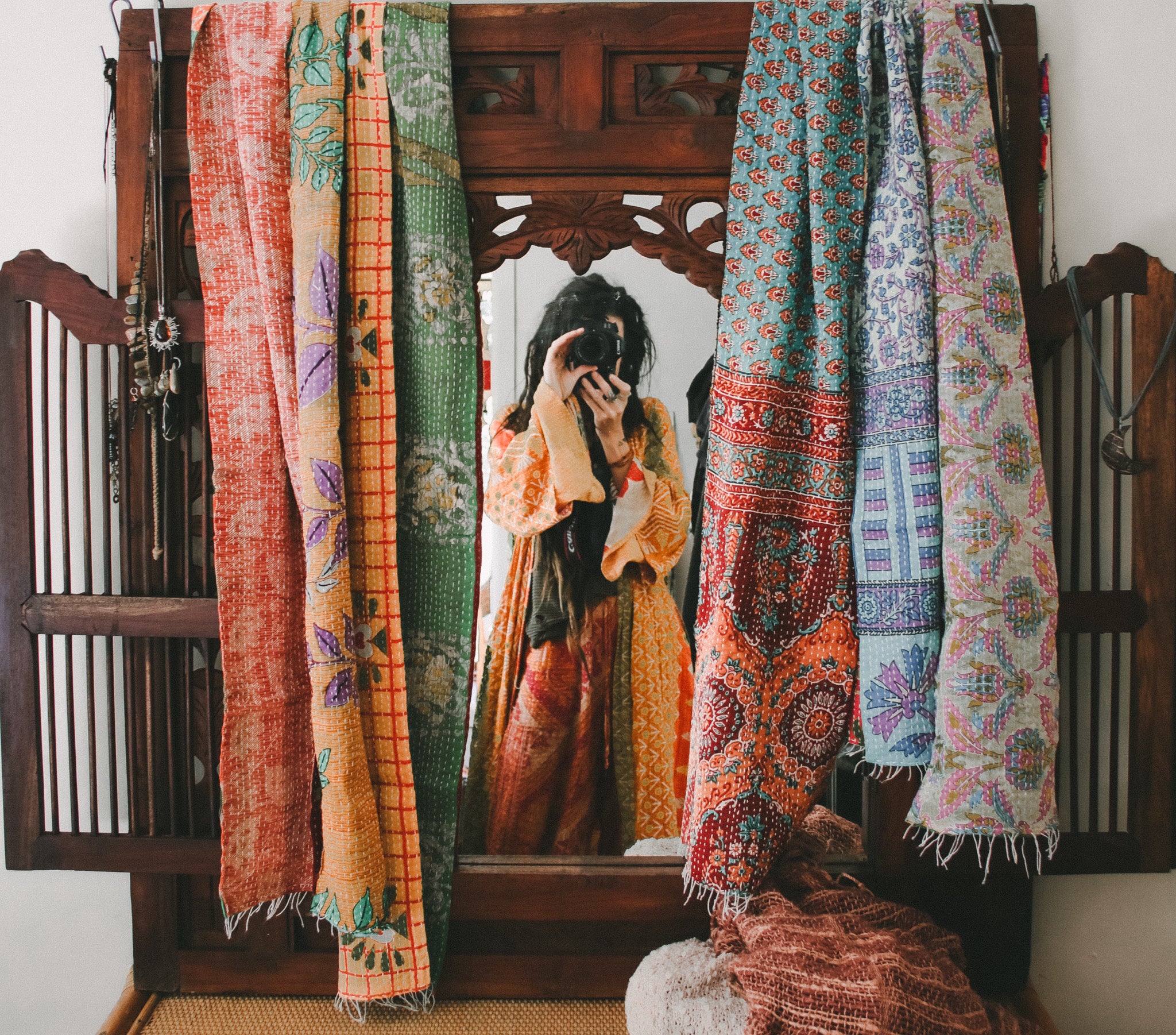 A mirror in a rustic wooden frame shows a person taking a photo, framed by several colorful patterned scarves hanging on both sides. The scarves feature intricate bohemian designs in orange, green, blue, and red.
