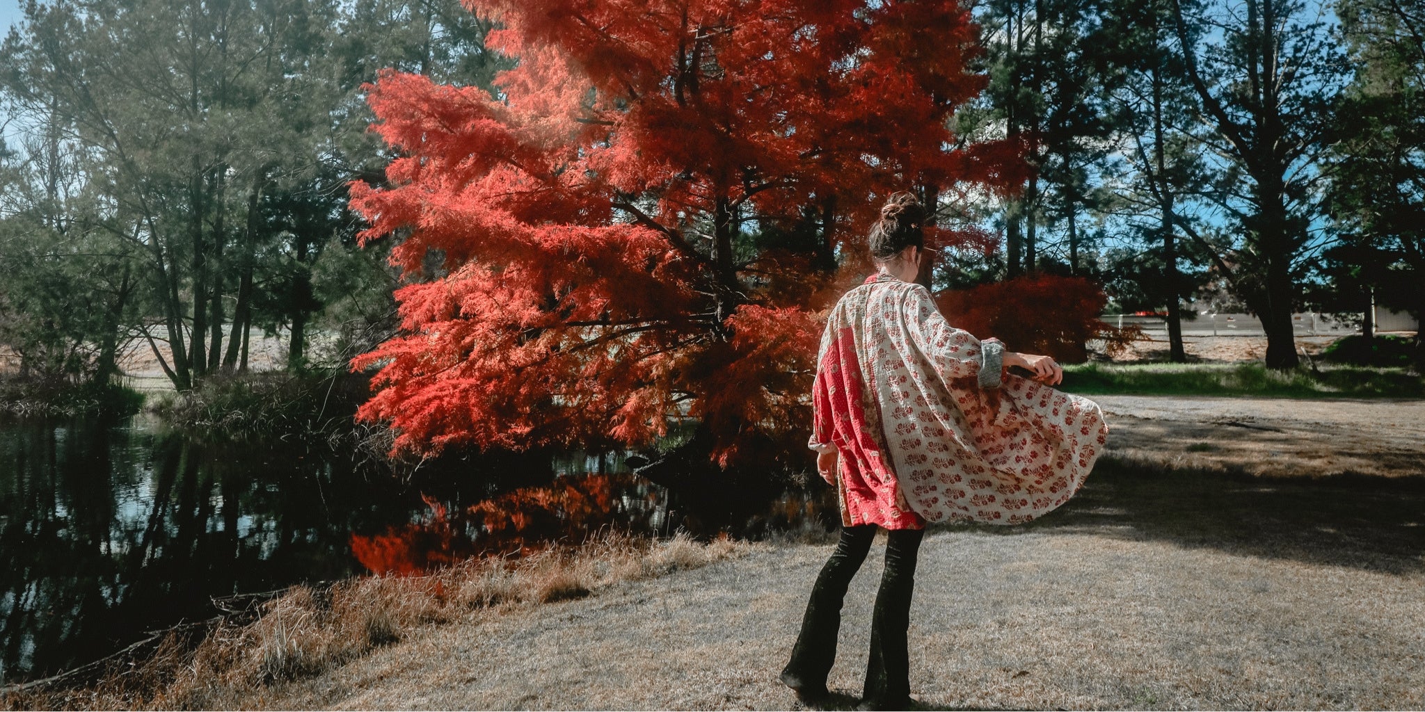 Person in a patterned dress standing near a tree with vibrant red leaves by a lake.