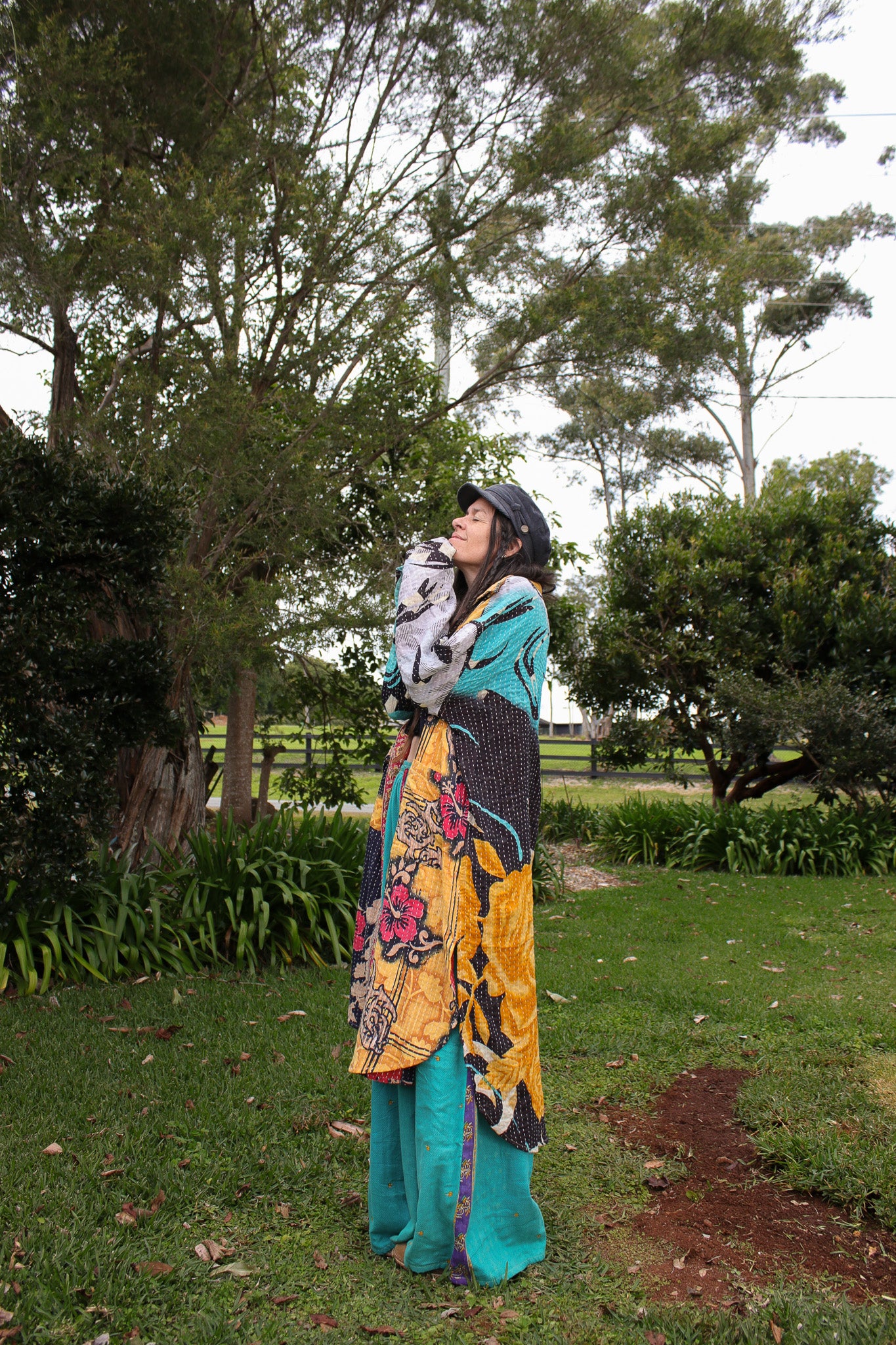A woman standing outdoors in a grassy park area wearing a long, colorful patchwork robe with floral patterns, teal pants, and a dark helmet, holding a light-colored cat.