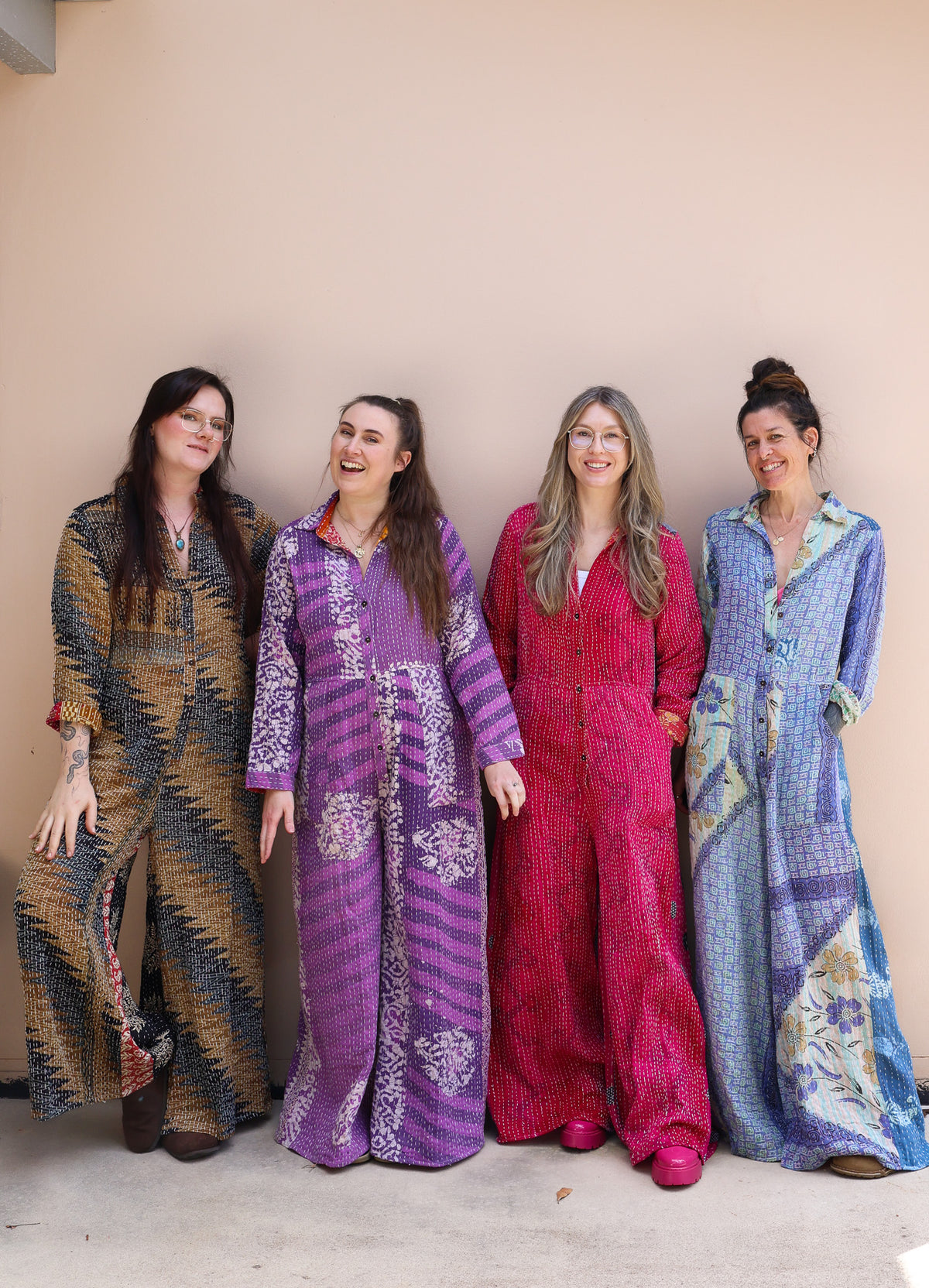 Four women standing side-by-side wearing matching long kaftan-style jumpsuits in different colorways: brown and black, purple and white, magenta and white, and blue and white. They are posing against a plain beige wall.