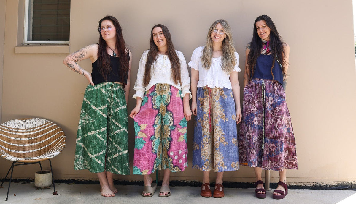 Four women standing outdoors against a beige stucco wall, each wearing a long patterned skirt paired with different tops. The skirts feature colorful prints including green, pink, blue, and purple, with bohemian-style patterns.