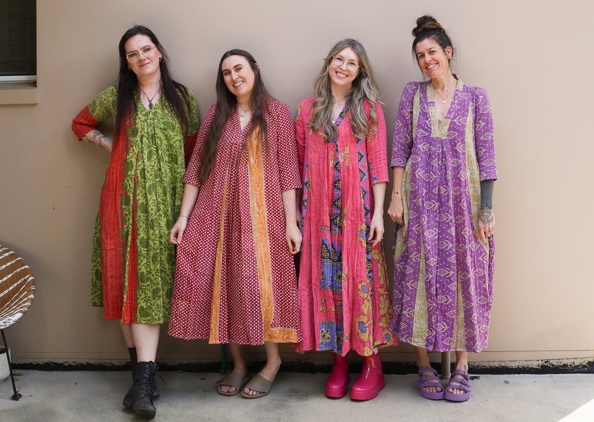Four women standing outdoors against a beige stucco wall, each wearing a long patterned kaftan dress in different colors including green, pink, red, and purple.