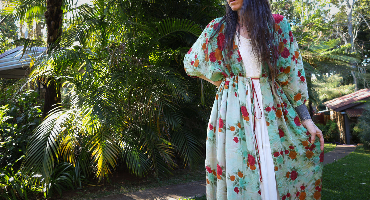 A woman wearing a long, flowing floral kimono-style dress over a white top, standing outdoors in a lush tropical garden setting.