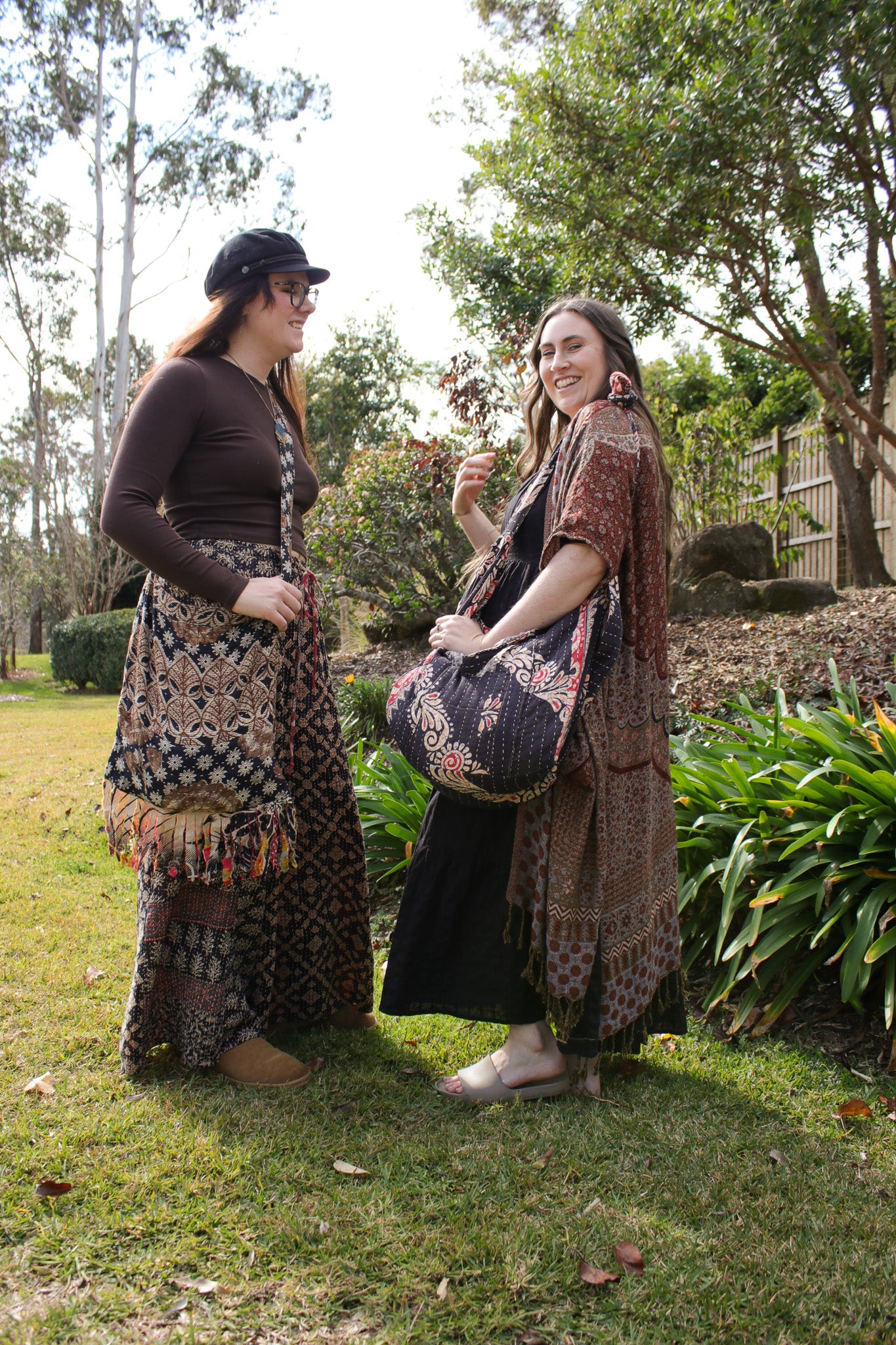 Two women standing outdoors on a grassy lawn wearing bohemian-style patterned clothing and carrying a matching patterned tote bag.